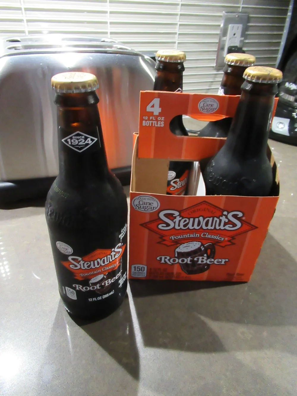Stewart's-Root-Beer-Glasflasche mit Goldkronkorken vor dem orangefarbenen Viererträger, Aufdruck „Since 1924“ und „Made with Cane Sugar“